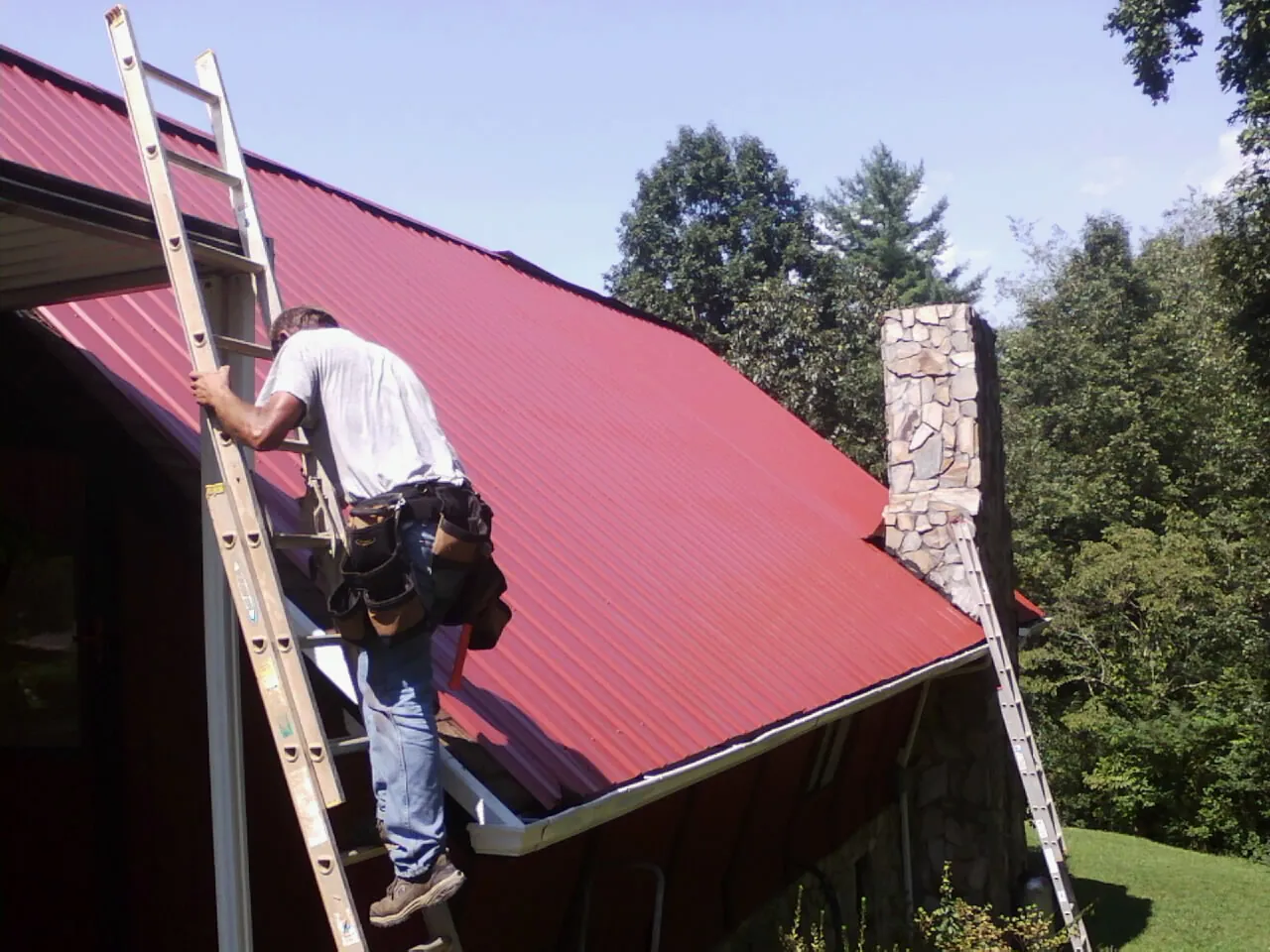 Roofer on a ladder beside a red metal roof and stone chimney — Soffit & Fascia Repair by Roofing Brothers in Hiram